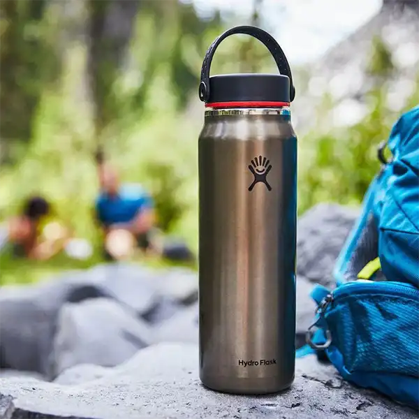 قمقمه آب Hydro Flask Lightweight Wide Mouth Trail بر روی یک صخره در کنار یک کوله پشتی با نمای پشت تیمه جنگلی سرسبز