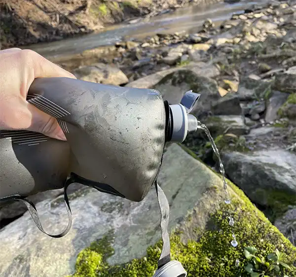 بطری آب HydraPak Stow 500 ml  در دست طبیعت گرد در حال فشار دادن و ریختن آب از ات با بکگراند ناحیه کوهستانی